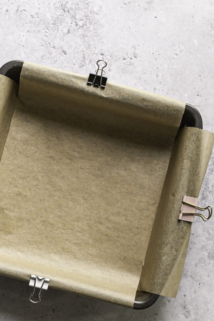 a close up of a square 8x8 baking pan lined with parchment paper and metal binder clips holding the parchment paper in place.