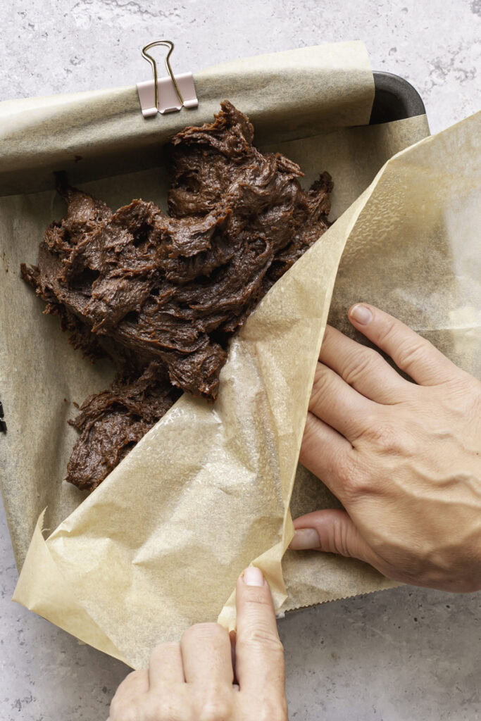 a hand pressing the brownie batter into a baking pan with a piece of parchment paper.