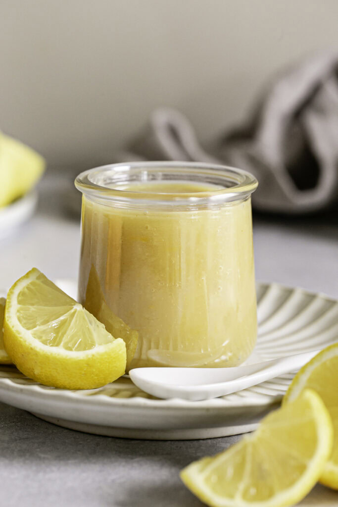 a close up of a jar of lemon curd on a small plate with a spoon beside it and slices of lemon surrounding it.