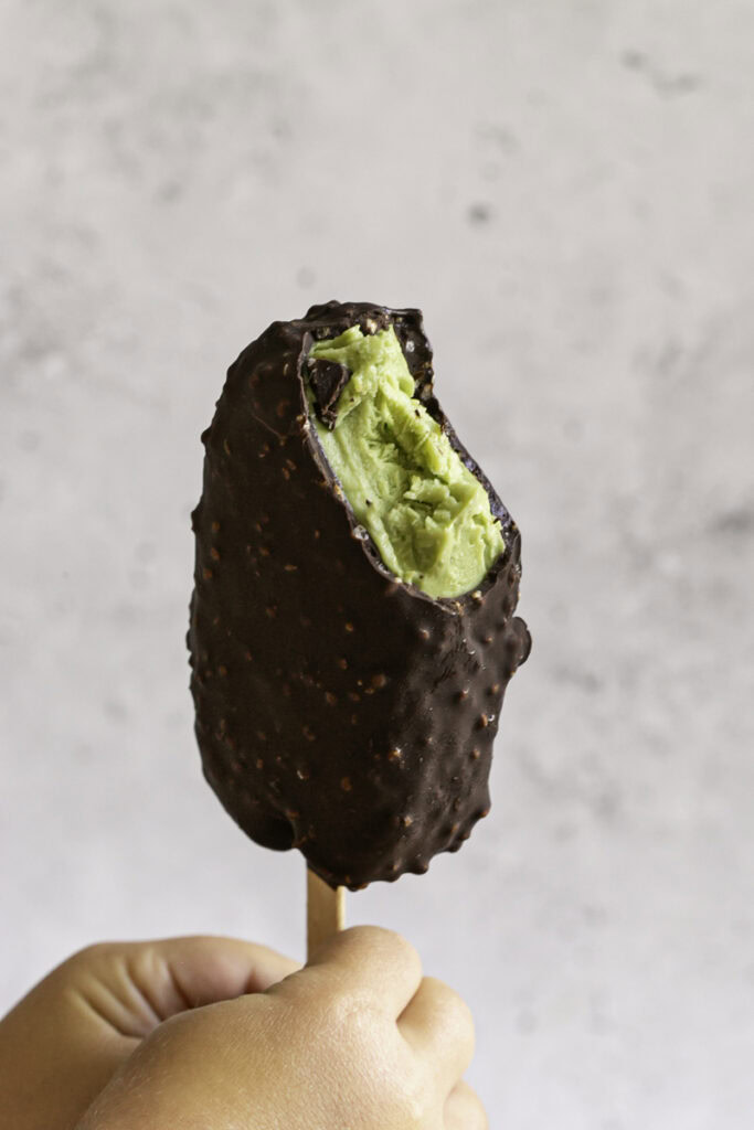 hands holding single creamy avocado mint chocolate ice cream bar with a bite taken out of it.