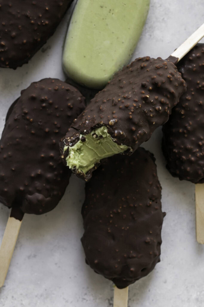 creamy avocado mint chocolate ice cream bars piled on top of one another, one not coated in chocolate and the top one with a bite taken out of it.