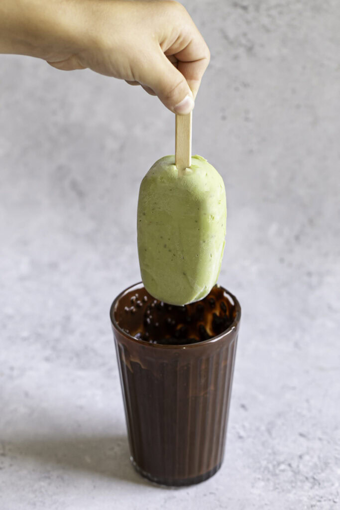 a hand holding the creamy avocado mint chocolate ice cream bar above the glass with melted chocolate.