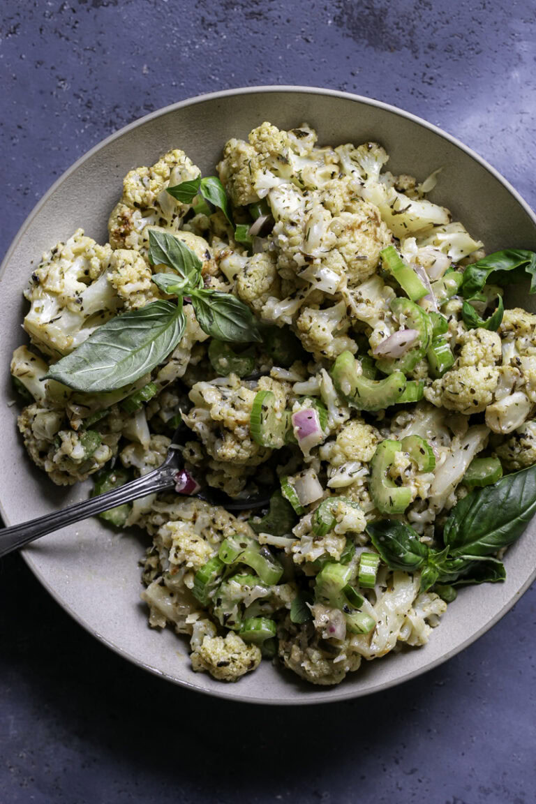 a bowl of Low Carb Roasted Cauliflower Potato Salad with a spoon sticking out of the bowl on a blue backdrop.