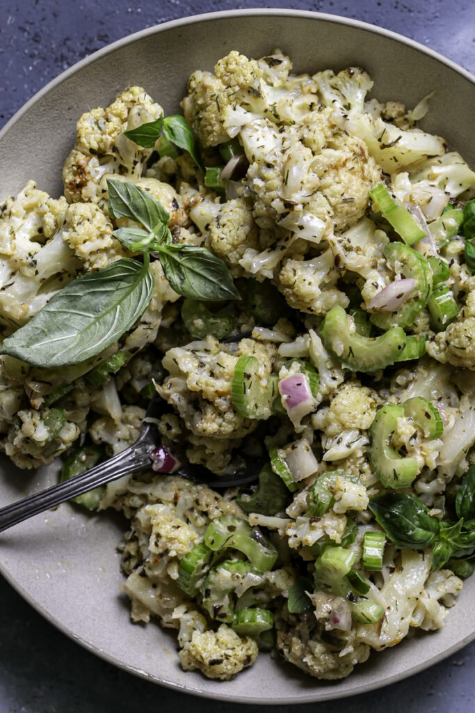 a close up of a bowl of Low Carb Roasted Cauliflower Potato Salad with a spoon sticking out of the bowl on a blue backdrop.