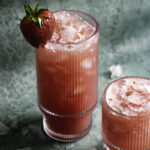 a tall glass of watermelon strawberry water over ice with a fresh strawberry on the rim of the glass, and a smaller glass of watermelon strawberry water over ice.