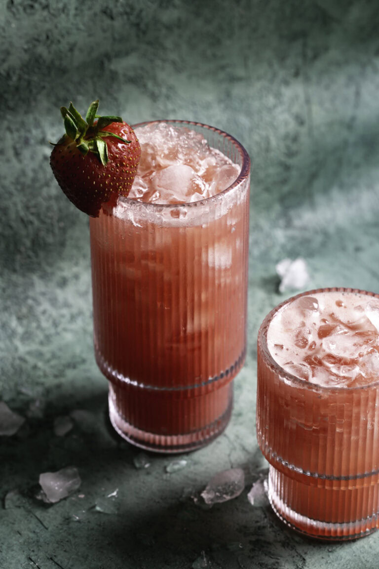 a tall glass of watermelon strawberry water over ice with a fresh strawberry on the rim of the glass, and a smaller glass of watermelon strawberry water over ice.