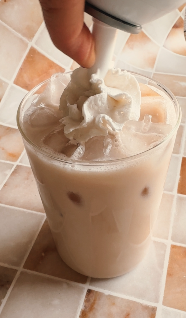 Action shot putting whipped cream on top of the iced chai latte.