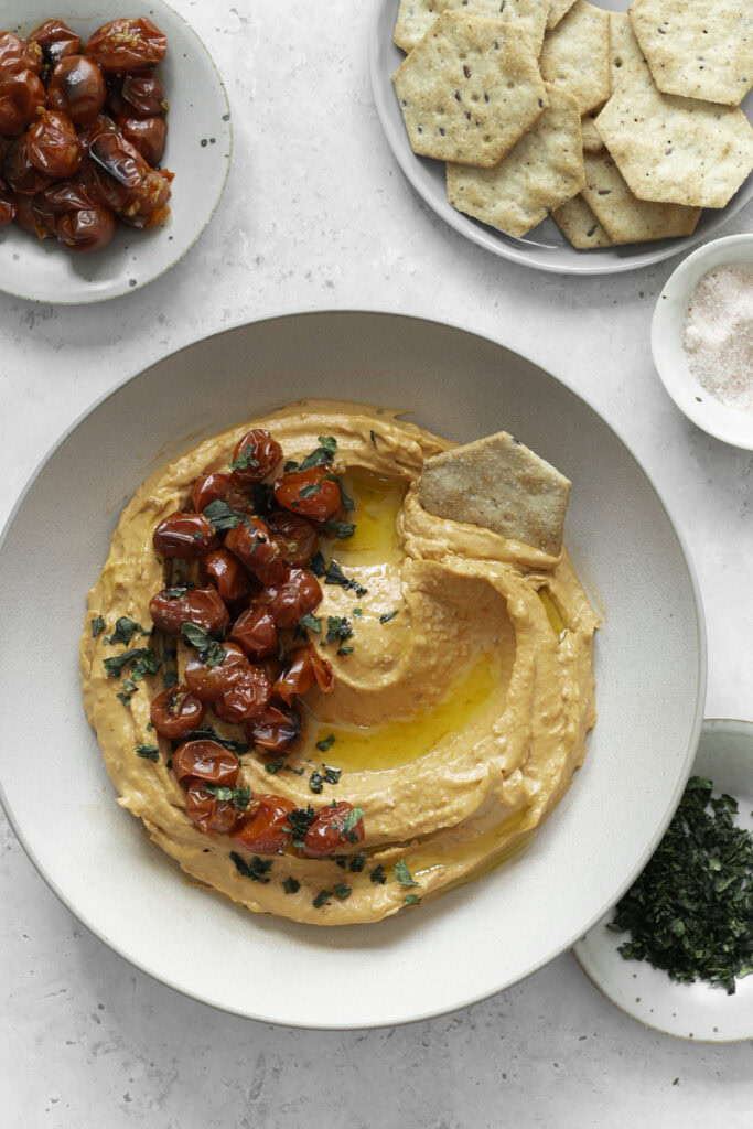 a bowl of roasted cherry tomato hummus with a cracker in it, a small plate of crackers above it, a small plate of roasted cherry tomatoes and chopped basil, and a small bowl of salt.