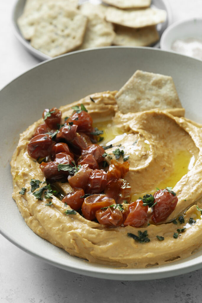 3/4 image of a bowl of roasted cherry tomato hummus garnished with roasted cherry tomatoes and a plate of crackers in the background.