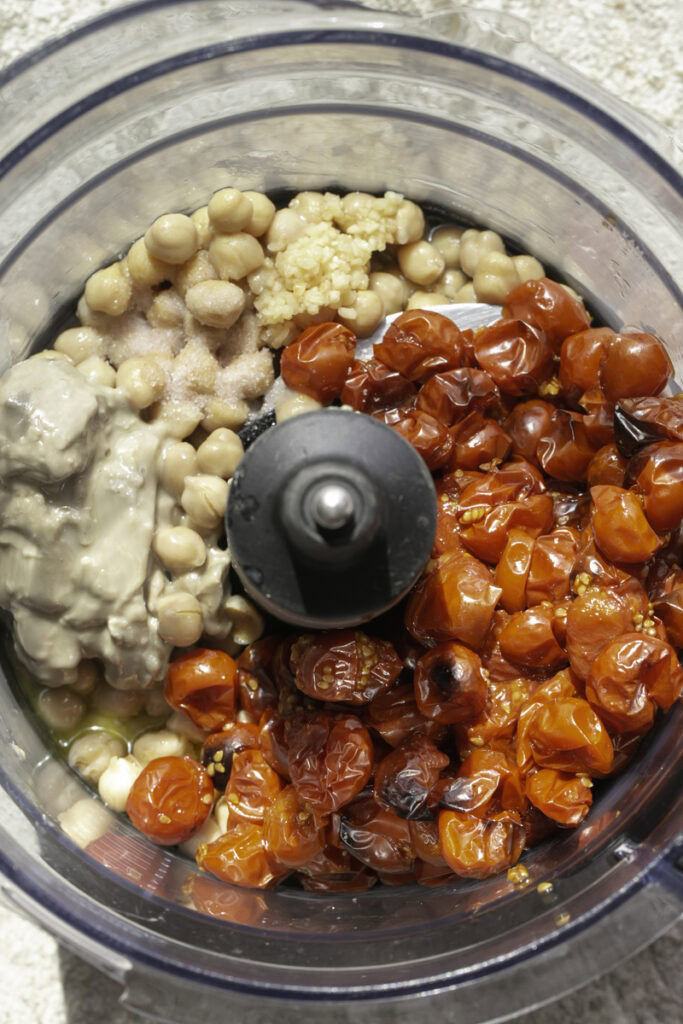 all of the ingredients to make roasted cherry tomato hummus in a food processor.