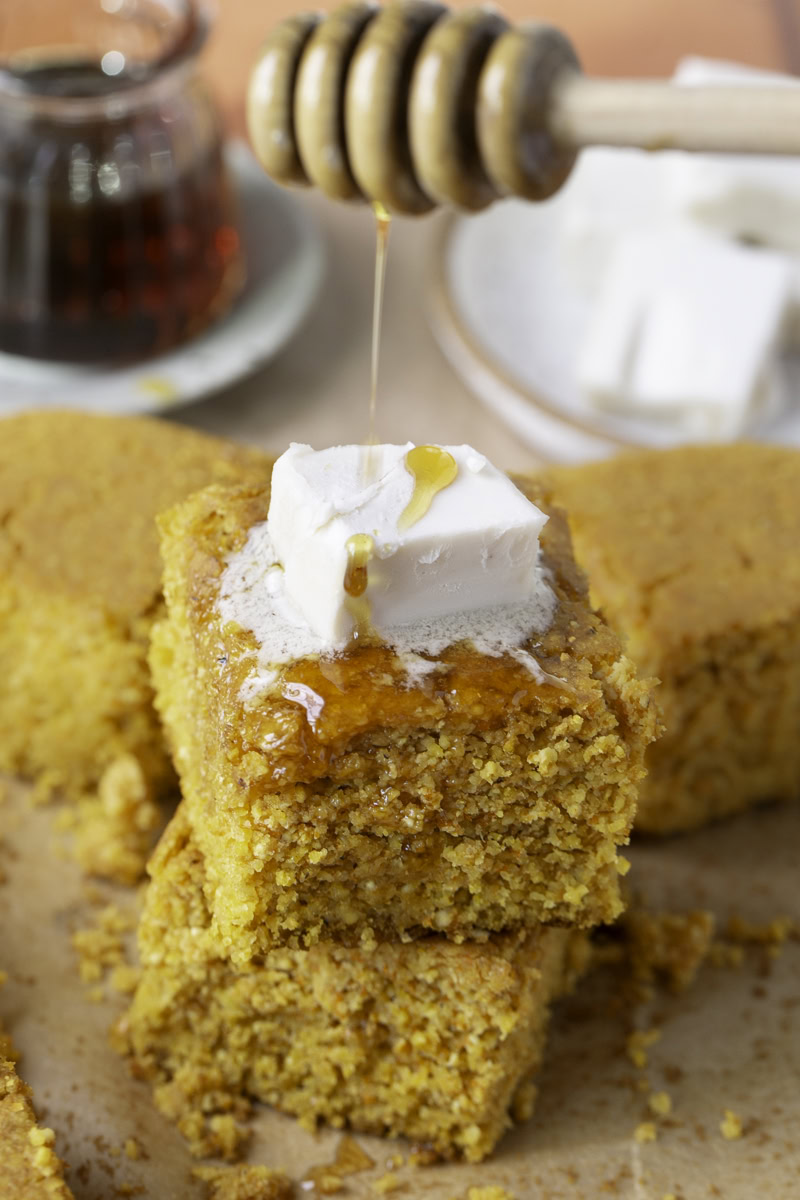 action shot drizzling maple syrup over a slice of cornbread topped with butter.