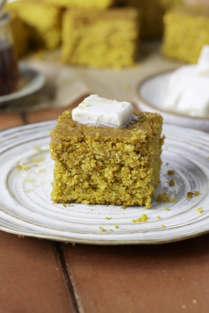 a slice of maple pumpkin cornbread on a plate topped with butter and other slices of cornbread in the background with a jar of maple syrup and a plate of butter.