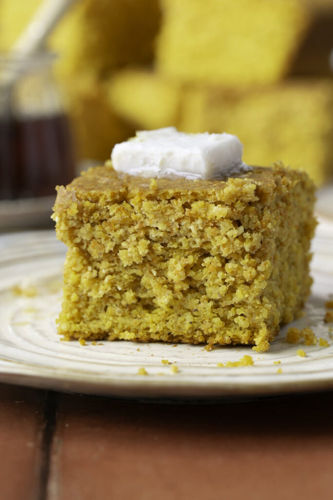 a slice of maple pumpkin cornbread on a plate topped with butter and other slices of cornbread in the background with a jar of maple syrup and a plate of butter.