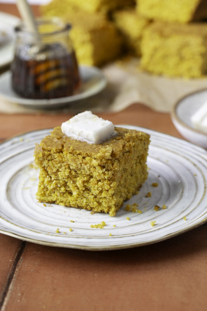 a slice of maple pumpkin cornbread on a plate topped with butter and other slices of cornbread in the background with a jar of maple syrup and a plate of butter.