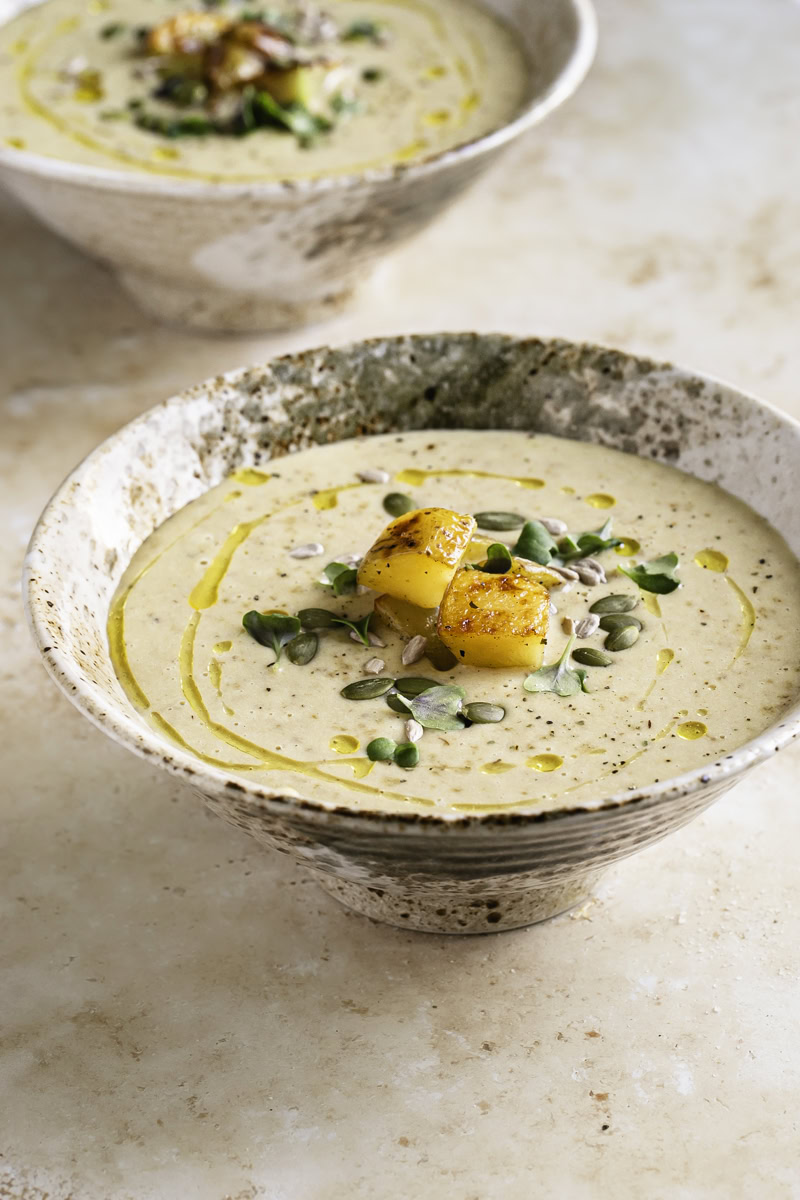 3/4 shot of 2 bowls of Creamy Roasted Gold Potato Leek Soup garnished with roasted potatoes, pumpkin and sunflower seeds and microgreens.