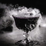 a close up of a black apple juice mocktail with dry ice smoking out of the glass.