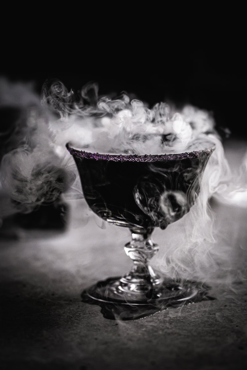 a close up of a black apple juice mocktail with dry ice smoking out of the glass.