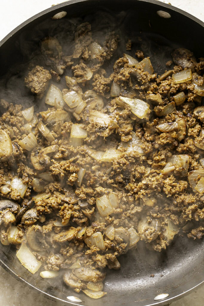 action shot cooking plant-based ground beef in a skillet with onions and mushrooms, with steam coming off the skillet.