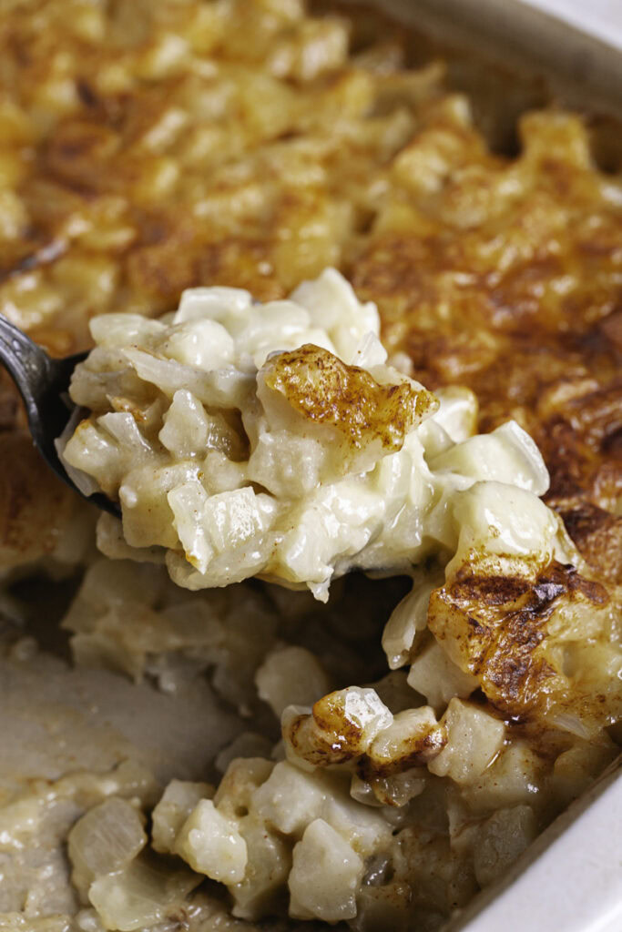 a casserole dish with baked creamy vegan hashbrown potatoes and a spoon scooping some out.