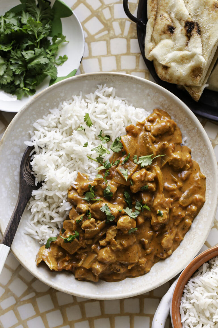 a bowl with basmati rice and indian butter chicken with a spoon in the bowl, a side bowl of rice and fresh cilantro leaves and a plate of naan.