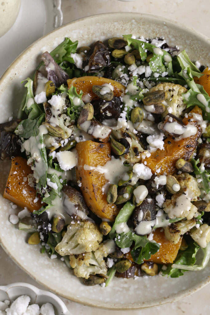 Roasted Butternut Squash Fall Harvest Salad with a jar of creamy apple cider vinaigrette, a bowl of pistachios, and a bowl of vegan feta cheese.