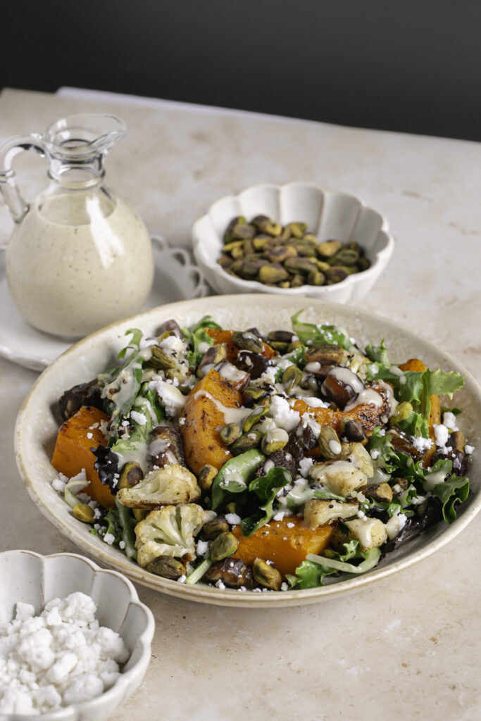 Roasted Butternut Squash Fall Harvest Salad with a jar of creamy apple cider vinaigrette, a bowl of pistachios, and a bowl of vegan feta cheese.