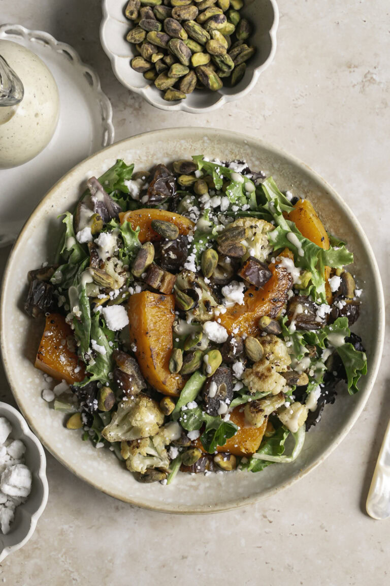Roasted Butternut Squash Fall Harvest Salad with a jar of creamy apple cider vinaigrette, a bowl of pistachios, and a bowl of vegan feta cheese.