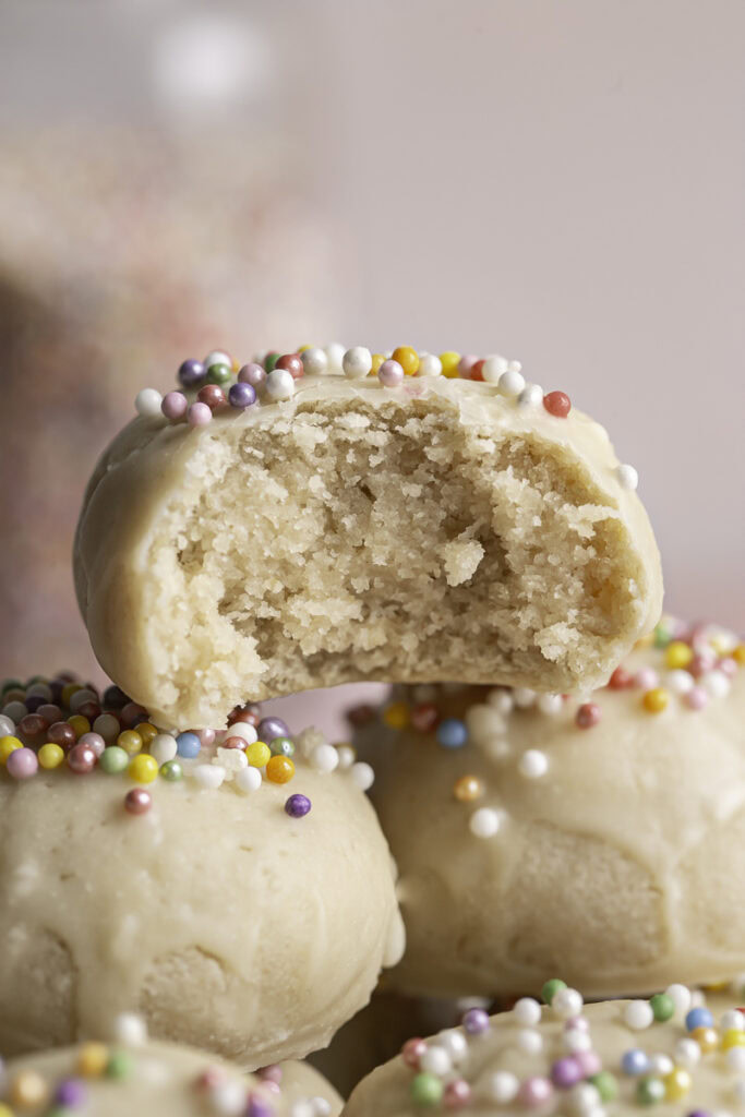 close up of Buttery Soft Italian Sprinkle Biscotti Cookies stacked on top of each other, the top one with a bite taken out of it to reveal the cookie texture.
