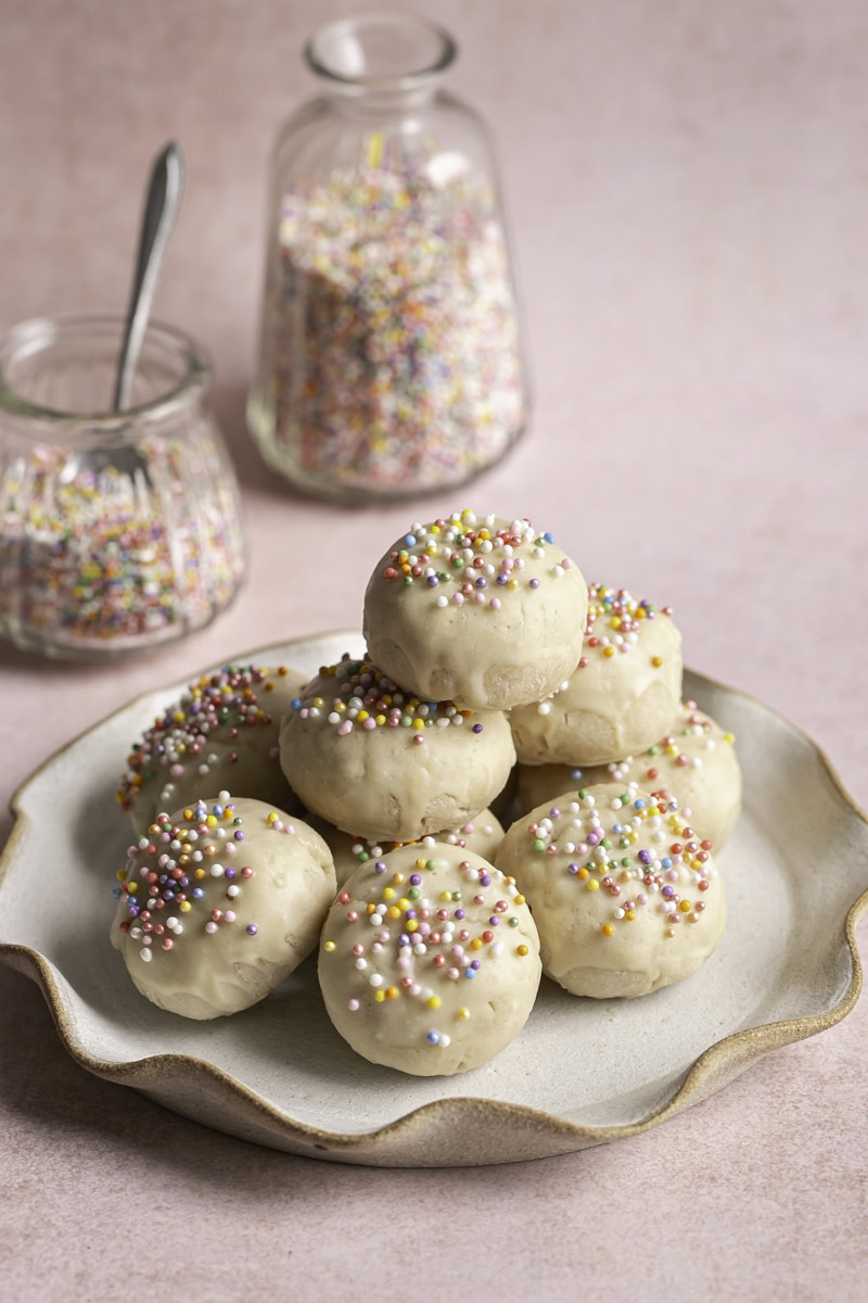 a plate of Buttery Soft Italian Sprinkle Biscotti Cookies with two glass jars of rainbow sprinkles in the background.