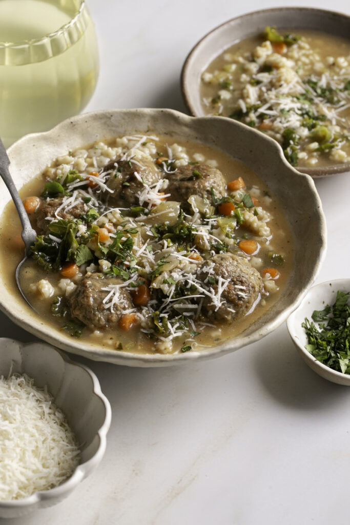 3/4 shot of a bowl of vegan italian wedding soup, a glass of white wine, a bowl of shredded vegan parmesan cheese, and a small bowl of soup.
