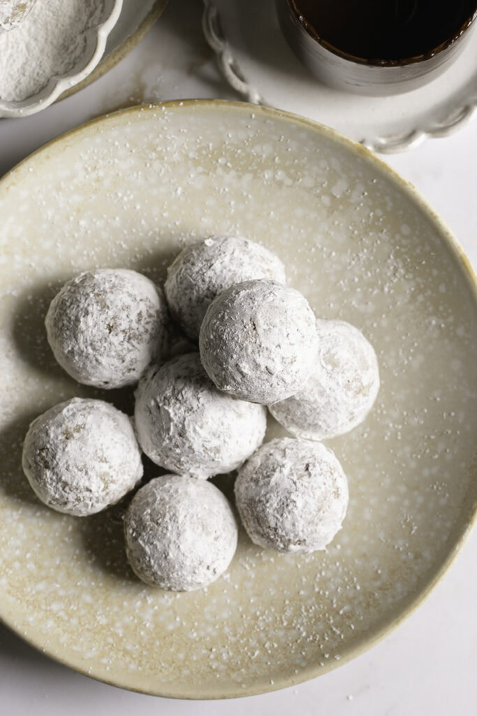 overhead shot of powdered sugar donut holes in a plate with a glass of melted chocolate and a small bowl of powdered sugar with a donut hole in it.