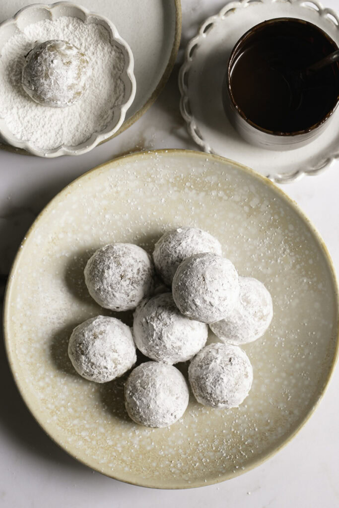 overhead shot of powdered sugar donut holes in a plate with a glass of melted chocolate and a small bowl of powdered sugar with a donut hole in it.