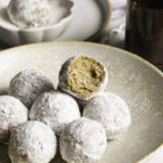 powdered sugar donut holes piled in a plate, the top one with a bite taken out of it and a glass of melted chocolate behind it.