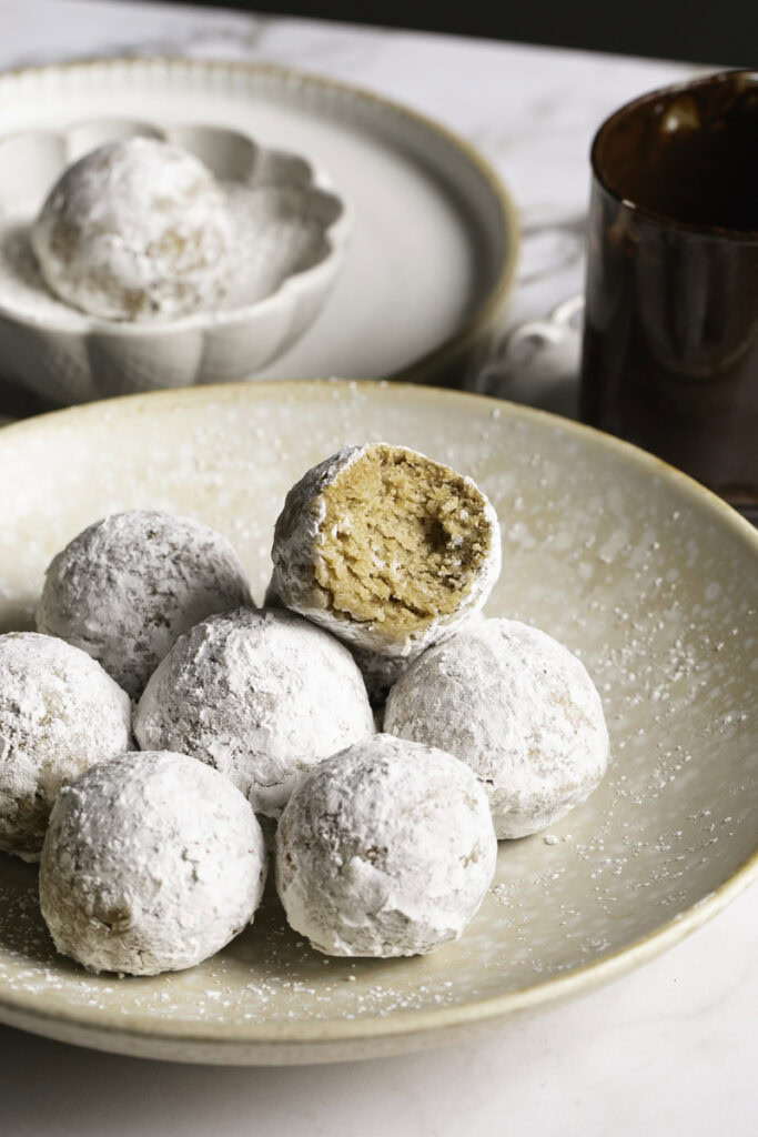 powdered sugar donut holes piled in a plate, the top one with a bite taken out of it and a glass of melted chocolate behind it.