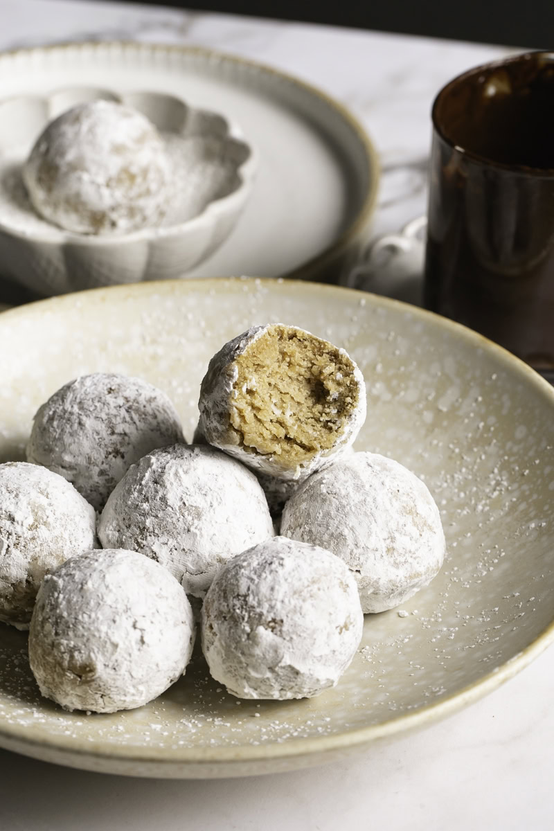 powdered sugar donut holes piled in a plate, the top one with a bite taken out of it and a glass of melted chocolate behind it.