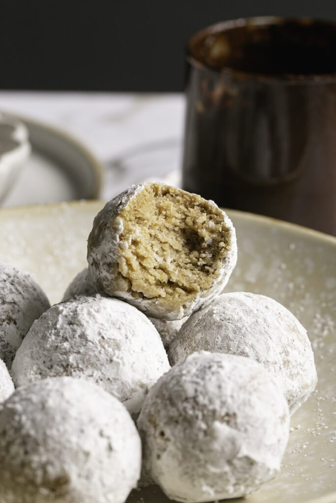 close up of powdered sugar donut holes piled in a plate, the top one with a bite taken out of it and a glass of melted chocolate behind it.