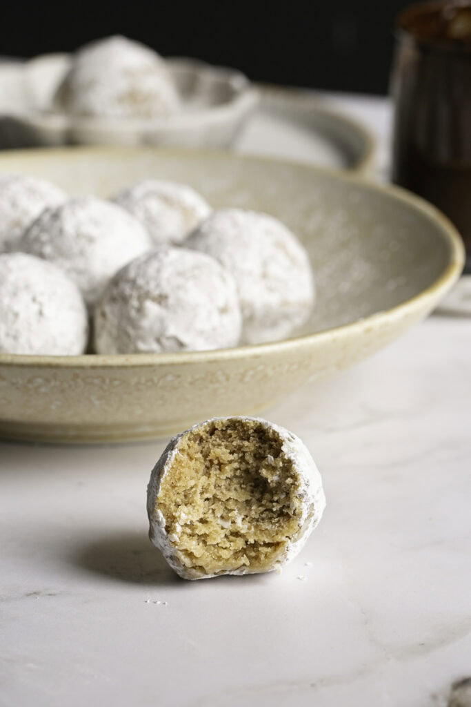 close up of a single donut hole with a bite taken out of it in front of a bowl of the other powdered sugar donut holes.