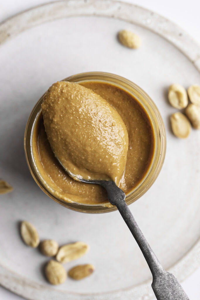 an overhead shot of homemade roasted peanut butter in a jar with a spoonful of peanut butter resting on the jar.