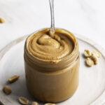 homemade roasted peanut butter in a glass jar with a spoon sticking out of it and whole roasted peanuts beside it.