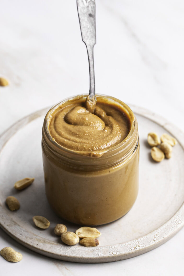 homemade roasted peanut butter in a glass jar with a spoon sticking out of it and whole roasted peanuts beside it.
