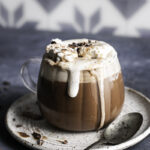 a glass of oat milk hot cocoa overflowing with whipped cream on a plate with a spoon.