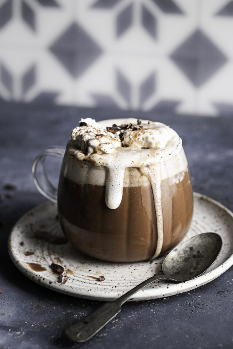 a glass of oat milk hot cocoa overflowing with whipped cream on a plate with a spoon.
