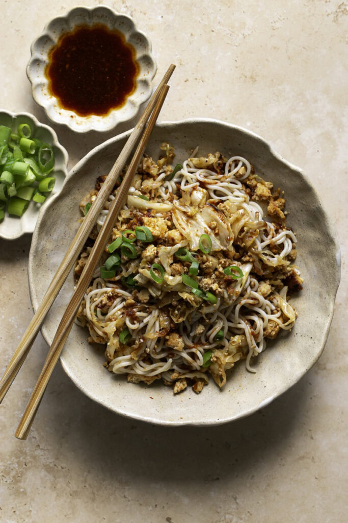 a bowl of Red Curry Tofu Cabbage Ramen Noodles with chopsticks, a small bowl of sliced green onions, and red curry soy sauce.