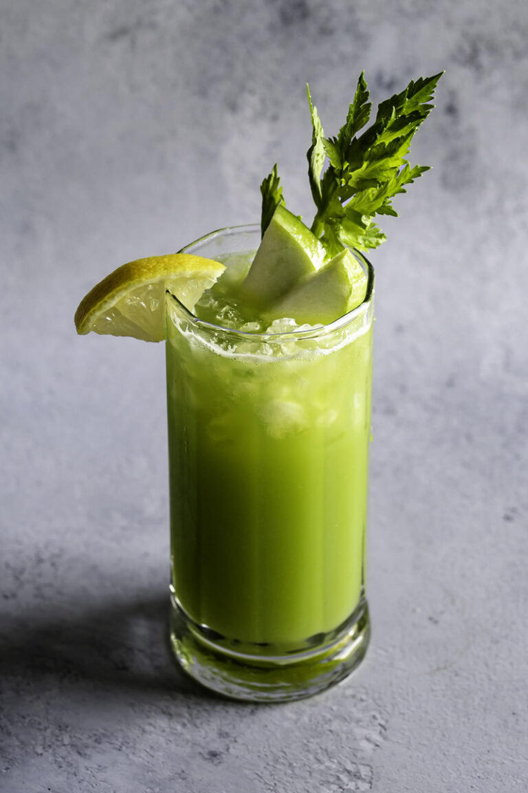a glass of green apple cucumber lemon celery juice garnished with slices of green apple, lemon, and celery.