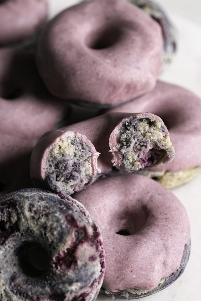 a close up of mini baked blackberry donuts, one donut without the frosting, and another donut with a bite taken out of it.