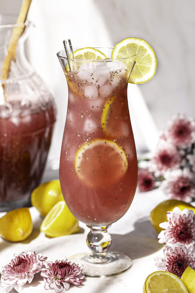 Pink Pitaya Chia Seed Lemonade in a glass with ice and lemon slices, fresh flowers around it and a pitcher of lemonade in the background.