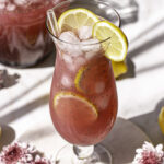 3/4 photo of Pink Pitaya Chia Seed Lemonade in a glass with ice and lemon slices, fresh flowers around it and a pitcher of lemonade in the background.