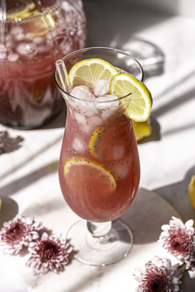 3/4 photo of Pink Pitaya Chia Seed Lemonade in a glass with ice and lemon slices, fresh flowers around it and a pitcher of lemonade in the background.