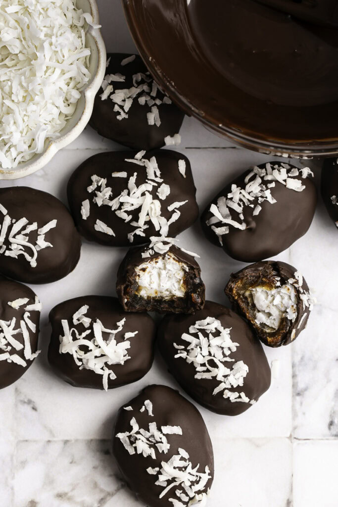 Vegan Bounty Bar Coconut Stuffed Dates in a pile, one with a bite taken out of it to reveal the coconut filling. A bowl of shredded coconut and a bowl of melted chocolate.