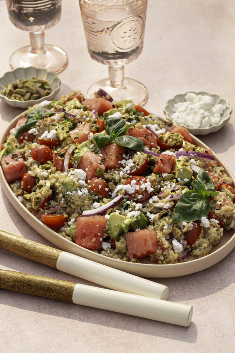 Watermelon Quinoa Salad on a serving plate with a bowl of feta cheese and a bowl of pistachios, two glasses of water, and salad servers.